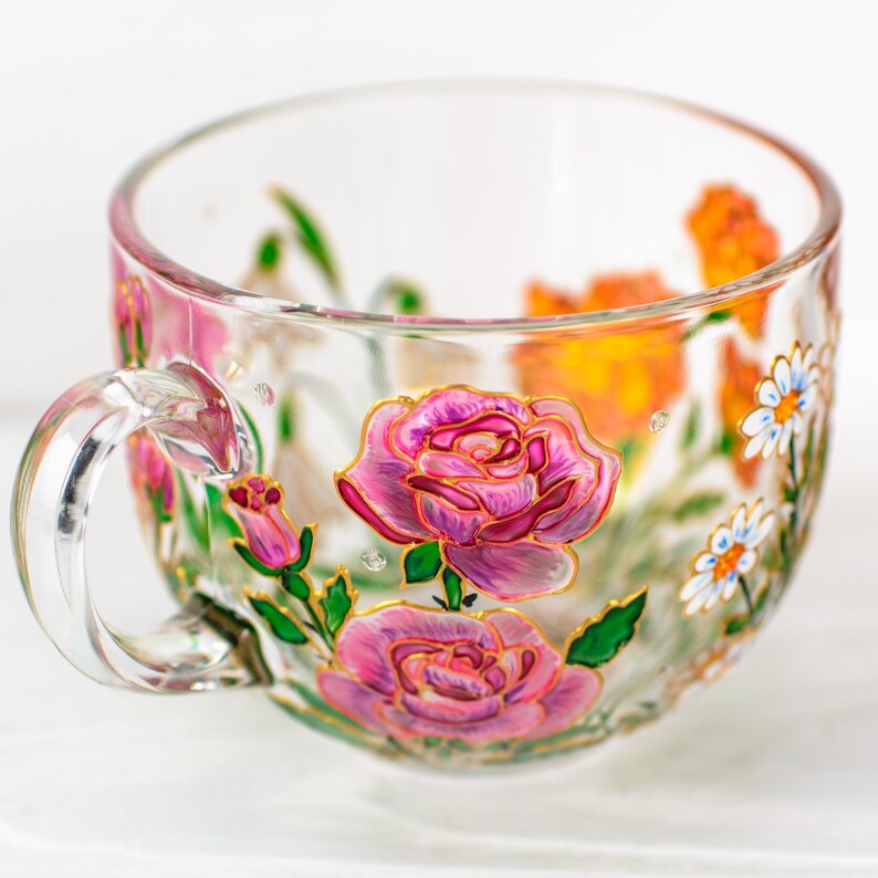 K&ouml;nnte beinhalten: Eine transparente Teetasse mit Henkel, handbemalt mit rosa Rosen, orangefarbenen Blumen und wei&szlig;en G&auml;nsebl&uuml;mchen. Das florale Design ist goldfarben umrandet, mit gr&uuml;nen Bl&auml;ttern und Stielen. Die Tasse steht auf einer wei&szlig;en Oberfl&auml;che.
