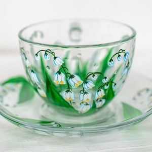 May include: A clear glass teacup and saucer set. The cup and saucer are decorated with hand-painted green leaves and white and blue lily of the valley flowers. The cup has a handle and the saucer is round.