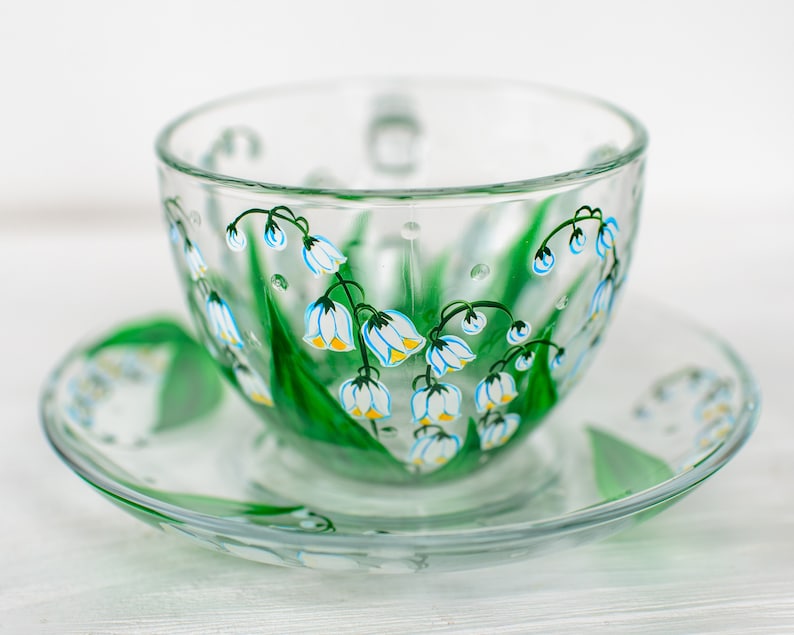 May include: A clear glass teacup and saucer set. The cup and saucer are decorated with hand-painted green leaves and white and blue lily of the valley flowers. The cup has a handle and the saucer is round.