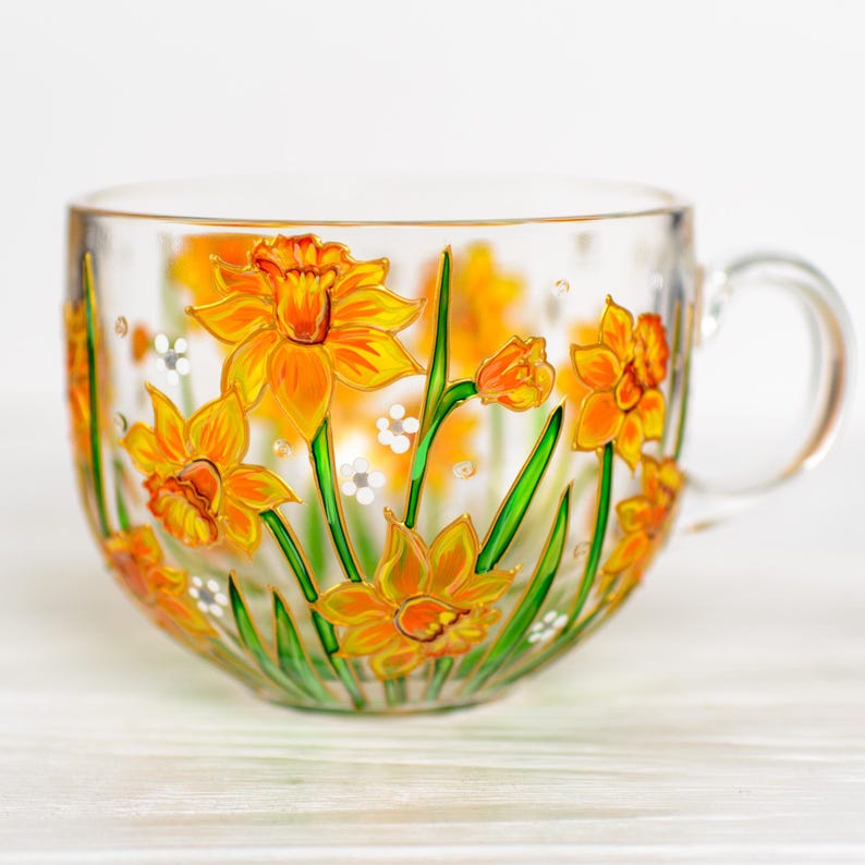 May include: A clear glass teacup with a handle, adorned with hand-painted yellow daffodils and green stems. The floral design includes white accents. The cup is ideal for serving tea or coffee.