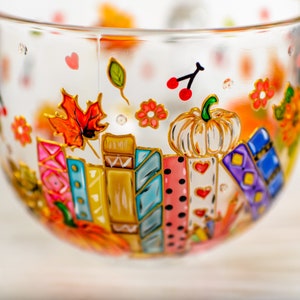Puede incluir: Una taza de cristal transparente decorada con dise&ntilde;os pintados a mano que muestran una fila de libros coloridos, una calabaza blanca, hojas de oto&ntilde;o, peque&ntilde;as flores rojas y corazones rojos. Las ilustraciones est&aacute;n delineadas en oro.