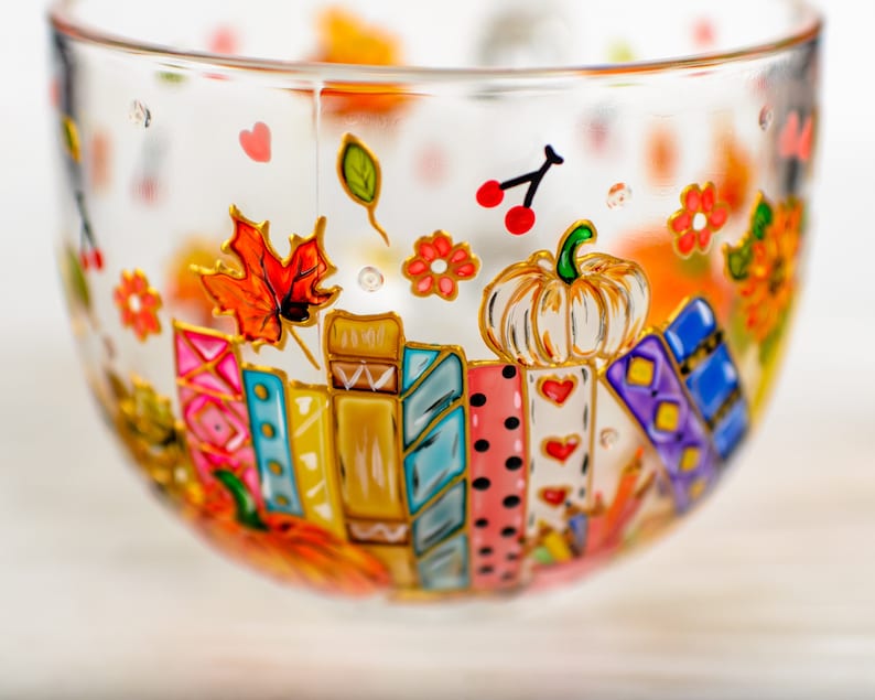 Puede incluir: Una taza de cristal transparente decorada con dise&ntilde;os pintados a mano que muestran una fila de libros coloridos, una calabaza blanca, hojas de oto&ntilde;o, peque&ntilde;as flores rojas y corazones rojos. Las ilustraciones est&aacute;n delineadas en oro.