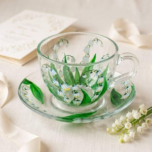 May include: A clear glass teacup and saucer set, hand-painted with a lily of the valley design. The cup and saucer feature green leaves and white bell-shaped flowers with yellow accents. A sprig of lily of the valley flowers is on the right.
