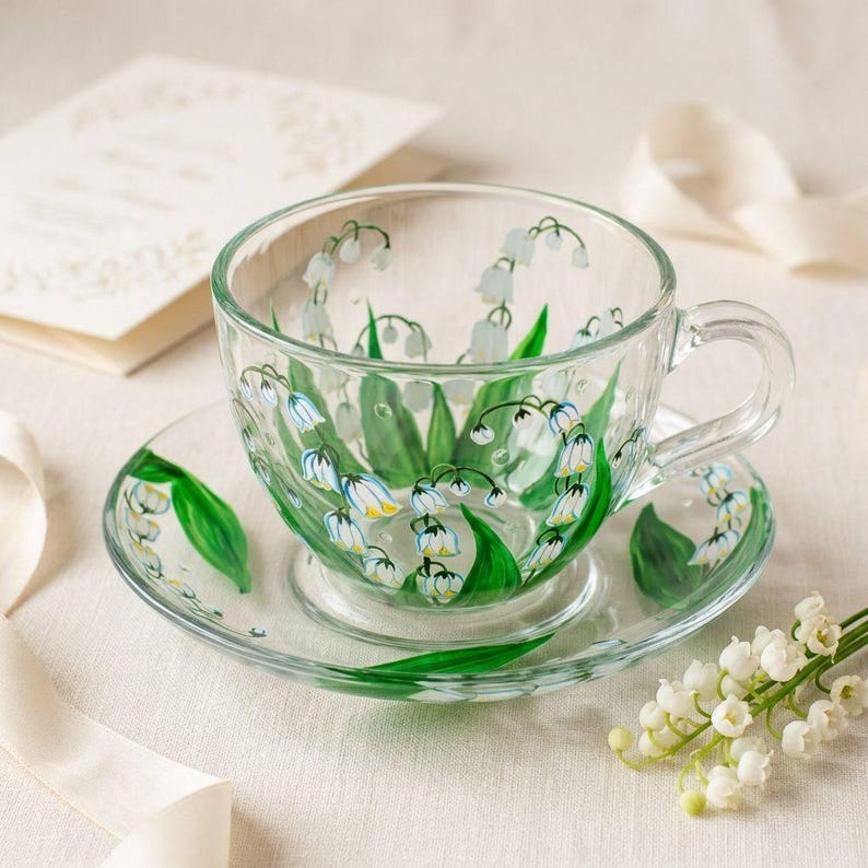 May include: A clear glass teacup and saucer set, hand-painted with a lily of the valley design. The cup and saucer feature green leaves and white bell-shaped flowers with yellow accents. A sprig of lily of the valley flowers is on the right.