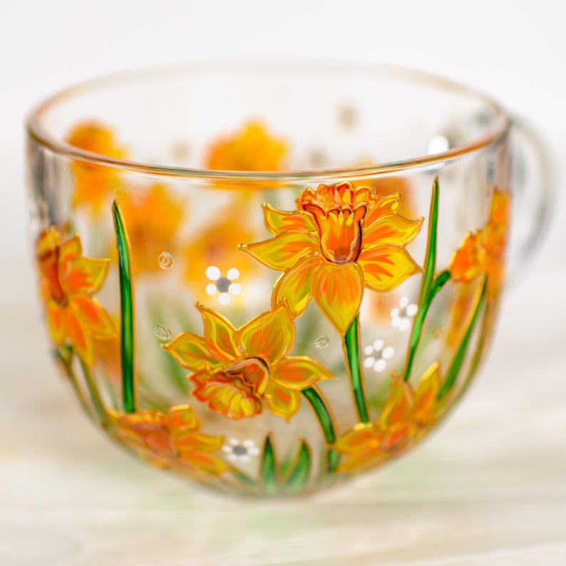 May include: A clear glass teacup with a handle, adorned with hand-painted yellow and orange daffodils and green stems. White daisy-like flowers are also painted on the cup. The cup is designed for tea or coffee.
