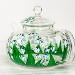 May include: A clear glass teapot with a painted design of white lily of the valley flowers with yellow centers and green leaves.