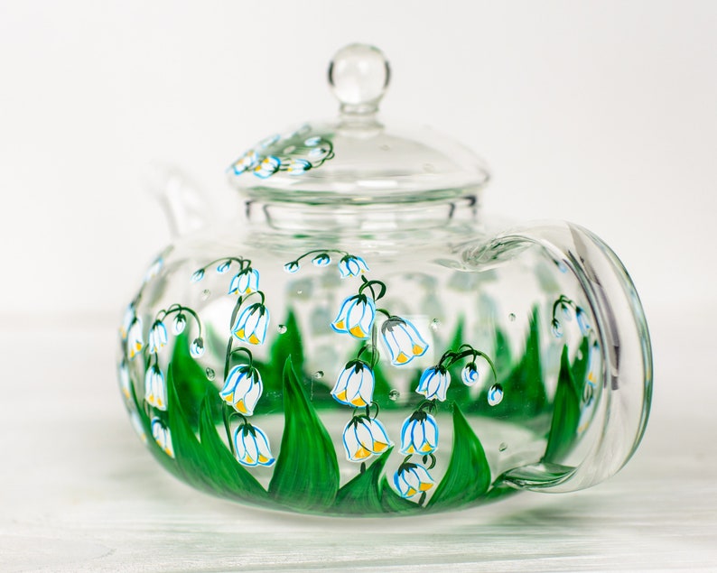 May include: A clear glass teapot with a painted design of white lily of the valley flowers with yellow centers and green leaves.