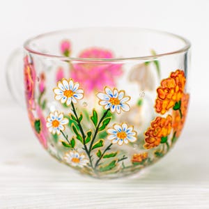 K&ouml;nnte beinhalten: Eine transparente Teetasse mit Henkel, handbemalt mit bunten Blumen. Wei&szlig;e G&auml;nsebl&uuml;mchen mit gelben Zentren und blauen Akzenten sind abgebildet, zusammen mit orangefarbenen und rosa Bl&uuml;ten. Das florale Design ist in Gold umrandet.