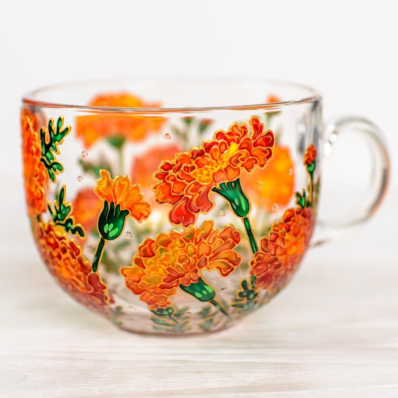 May include: A clear glass teacup with a large handle, adorned with hand-painted orange marigold flowers and green stems. The floral design wraps around the cup, creating a vibrant and artistic look. The cup is ideal for serving tea or other beverages.