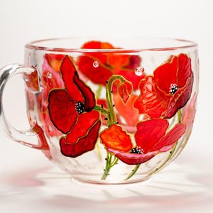Puede incluir: Una taza de t&eacute; de vidrio transparente con asa, adornada con amapolas rojas pintadas a mano. Las flores tienen centros negros y contornos dorados. La taza est&aacute; decorada con tonos rojos y naranjas.