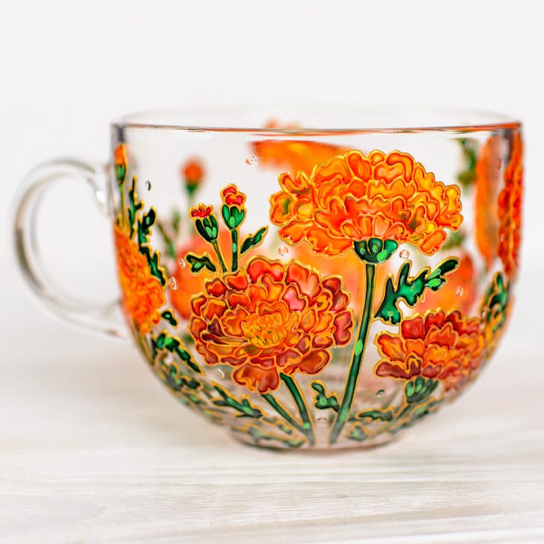 May include: A clear glass teacup with a handle, adorned with hand-painted orange marigold flowers and green stems. The floral design wraps around the cup, creating a vibrant and decorative piece. The cup is perfect for enjoying tea or other beverages.