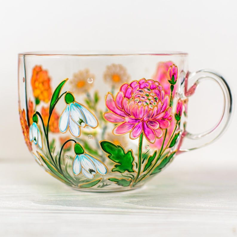 K&ouml;nnte beinhalten: Eine transparente Teetasse mit Henkel, verziert mit handgemalten Blumenmustern. Die Tasse zeigt eine gro&szlig;e rosa Chrysantheme, wei&szlig;e Schneegl&ouml;ckchen und orangefarbene Blumen, alle mit Gold umrandet. Gr&uuml;ne Bl&auml;tter und Stiele vervollst&auml;ndigen das botanische Thema.