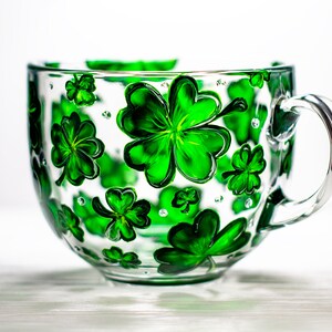 May include: A clear glass teacup with a large handle, decorated with hand-painted green shamrocks. The shamrocks vary in size and are scattered across the cup's surface. The cup is sitting on a white surface.