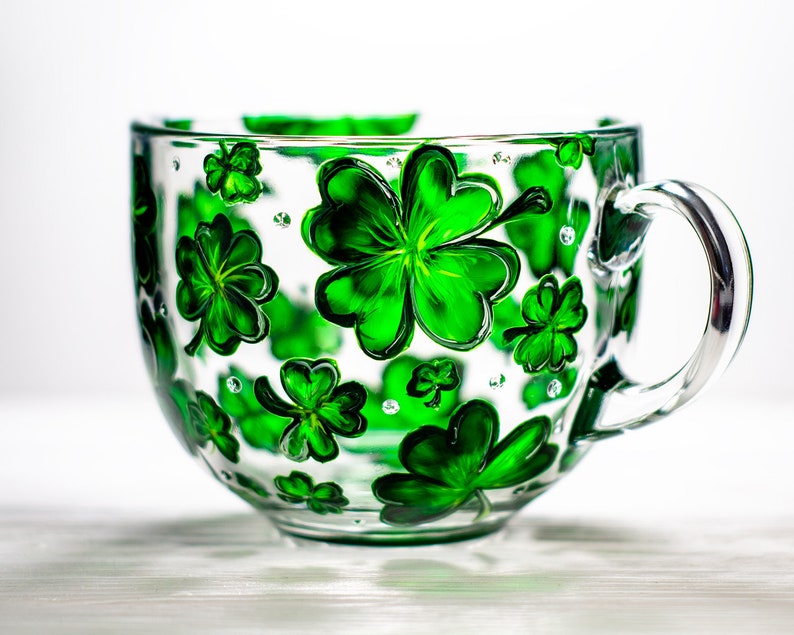 May include: A clear glass teacup with a large handle, decorated with hand-painted green shamrocks. The shamrocks vary in size and are scattered across the cup's surface. The cup is sitting on a white surface.
