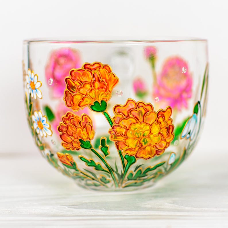 K&ouml;nnte beinhalten: Eine transparente Glasschale mit handgemalten Blumenmotiven. Sie zeigt leuchtend orange Ringelblumen, rosa Bl&uuml;ten und wei&szlig;e G&auml;nsebl&uuml;mchen mit goldenen Akzenten. Gr&uuml;ne Stiele und Bl&auml;tter erg&auml;nzen das botanische Thema.