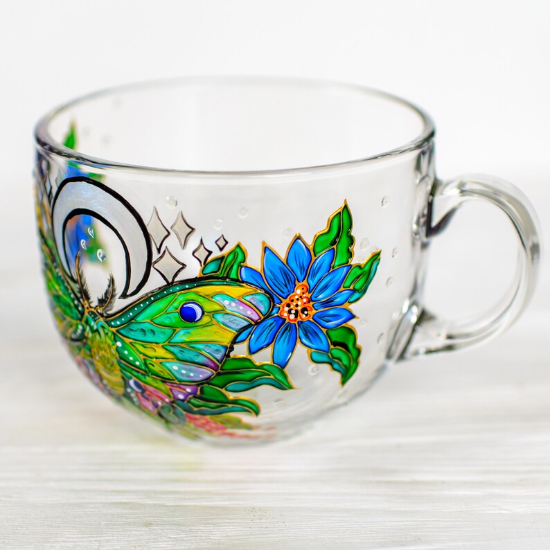 May include: A clear glass teacup with a handle, decorated with a colorful hand-painted design. The design features a blue flower, a green and yellow fish, and a crescent moon with stars. The cup is on a white wooden surface.