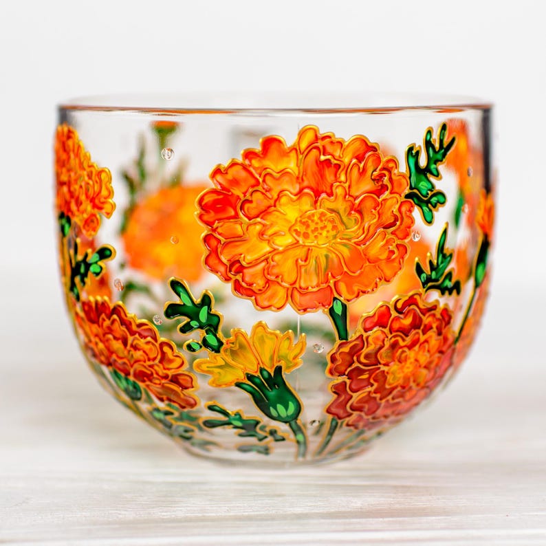 May include: A clear glass bowl decorated with vibrant orange and yellow marigold flowers. The flowers are detailed with gold outlines and green leaves, creating a colorful floral design. The bowl sits on a white surface.
