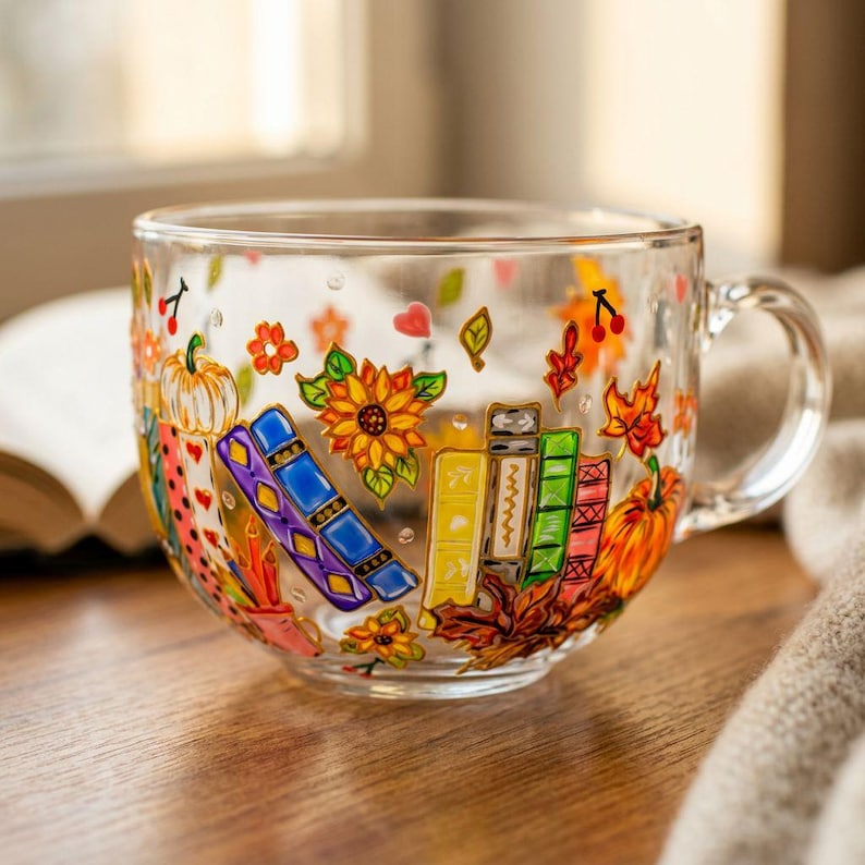 Puede incluir: Una taza de cristal transparente con un dise&ntilde;o pintado de colores que muestra libros apilados, calabazas, girasoles y hojas de oto&ntilde;o. La ilustraci&oacute;n incluye tonos vibrantes en naranja, azul, amarillo, verde y morado, con peque&ntilde;os corazones.