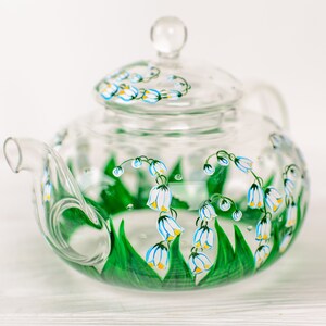 May include: A clear glass teapot with a lid, hand-painted with white and yellow lily of the valley flowers and green leaves.
