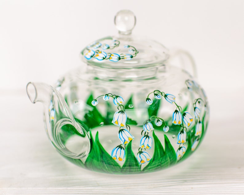 May include: A clear glass teapot with a lid, hand-painted with white and yellow lily of the valley flowers and green leaves.