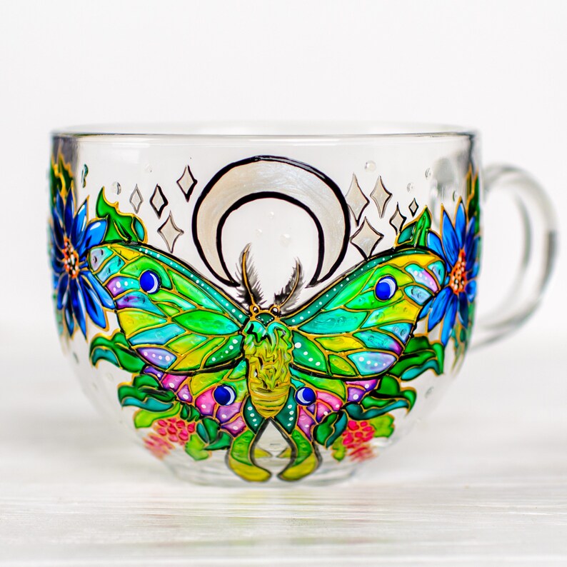 May include: A clear glass teacup with a hand-painted design featuring a green and yellow butterfly, blue flowers, and a silver crescent moon. The cup has a handle and is decorated with colorful details and sparkling accents.