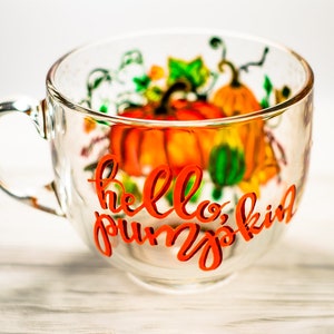 May include: A clear glass mug featuring a hand-painted design of orange pumpkins and green vines. The text "hello, pumpkin" is written in a bold, orange cursive script across the front of the mug, which has a single handle on the left side.