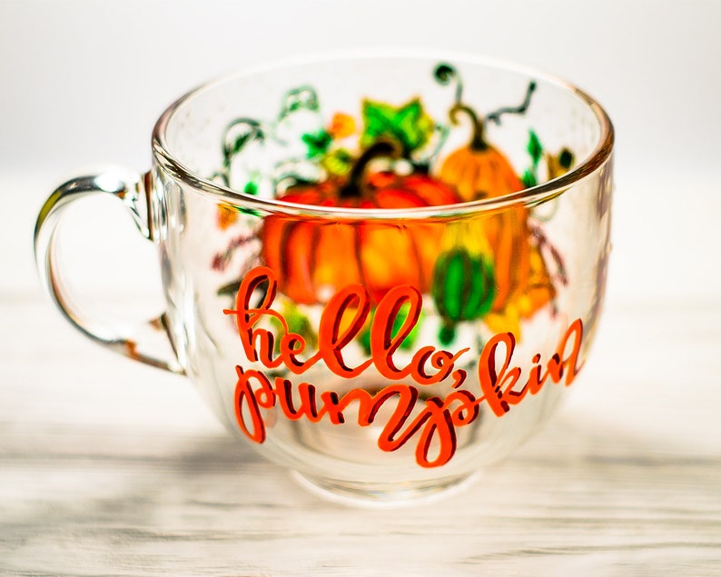 May include: A clear glass mug featuring a hand-painted design of orange pumpkins and green vines. The text "hello, pumpkin" is written in a bold, orange cursive script across the front of the mug, which has a single handle on the left side.