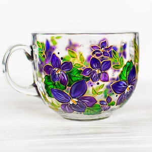 Puede incluir: Una taza de t&eacute; de vidrio transparente con asa, adornada con violetas moradas pintadas a mano, hojas verdes y detalles dorados. El dise&ntilde;o floral envuelve la taza, creando un aspecto decorativo y art&iacute;stico. La taza est&aacute; sobre un fondo blanco.