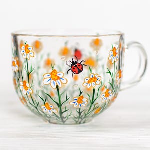 Puede incluir: Una taza de t&eacute; de vidrio transparente con un dise&ntilde;o floral pintado. La taza presenta margaritas blancas y amarillas, tallos verdes y una mariquita roja. El asa es transparente y redondeada. El dise&ntilde;o est&aacute; pintado en el exterior de la taza.