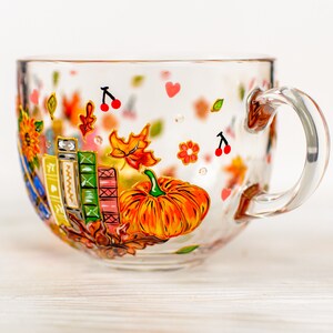 Puede incluir: Una taza de cristal transparente con dise&ntilde;os pintados a mano de una calabaza naranja brillante, una pila de libros coloridos, hojas de oto&ntilde;o, peque&ntilde;as cerezas rojas y diminutos corazones rosas. La taza tiene un asa curva y un cuerpo redondeado.