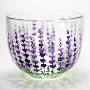 May include: A clear glass bowl featuring a hand-painted design of vertical lavender sprigs in various shades of purple and violet. The floral pattern wraps around the exterior of the rounded vessel, showcasing detailed petals and green stems.