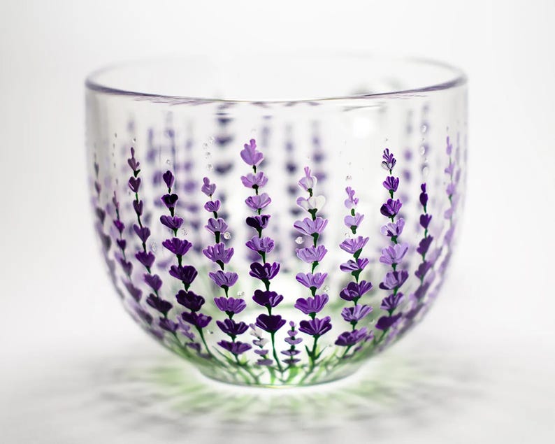 May include: A clear glass bowl featuring a hand-painted design of vertical lavender sprigs in various shades of purple and violet. The floral pattern wraps around the exterior of the rounded vessel, showcasing detailed petals and green stems.