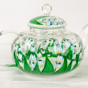 May include: A clear glass teapot with a painted design of white and blue lily of the valley flowers with green leaves.