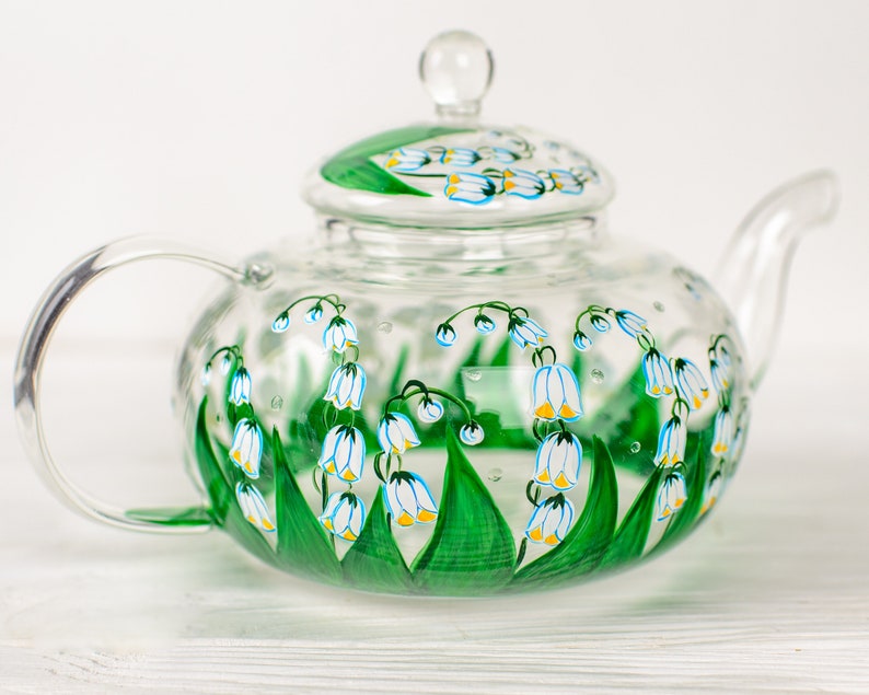 May include: A clear glass teapot with a painted design of white and blue lily of the valley flowers with green leaves.
