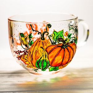 May include: A clear glass mug featuring a hand-painted design of vibrant orange pumpkins, a small green gourd, and green leaves. The artwork includes fine black outlines and delicate details, with a handle on the right side and a smooth, transparent finish.