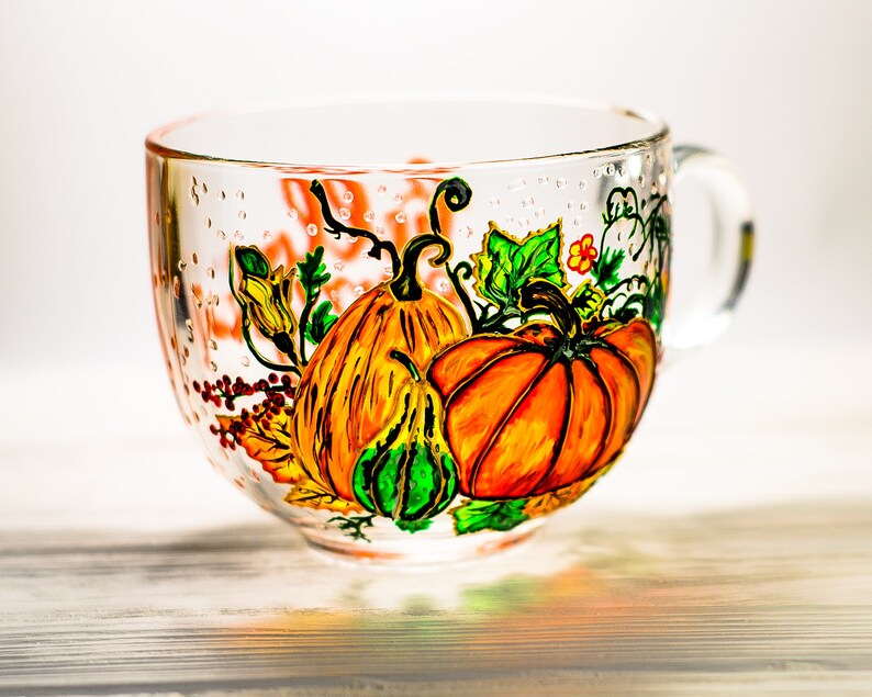 May include: A clear glass mug featuring a hand-painted design of vibrant orange pumpkins, a small green gourd, and green leaves. The artwork includes fine black outlines and delicate details, with a handle on the right side and a smooth, transparent finish.