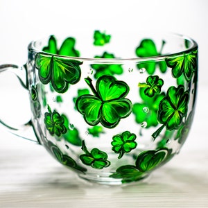 May include: A clear glass mug with a large handle, decorated with hand-painted green shamrocks. The shamrocks vary in size and are scattered across the surface of the mug. The mug is ideal for serving beverages.