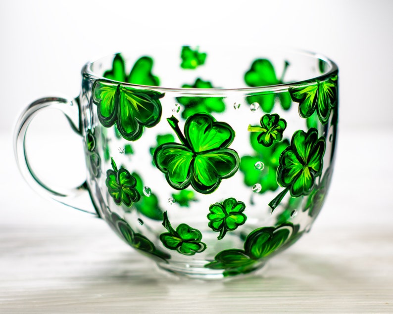 May include: A clear glass mug with a large handle, decorated with hand-painted green shamrocks. The shamrocks vary in size and are scattered across the surface of the mug. The mug is ideal for serving beverages.