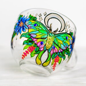 May include: A clear glass teacup with a hand-painted design featuring a colorful moth with green, yellow, blue, and pink wings. The cup also has blue flowers, pink berries, and a crescent moon design.