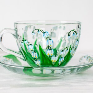 May include: A clear glass teacup and saucer set. The cup and saucer are decorated with hand-painted lily of the valley flowers in white, blue, and green. The cup has a large handle and sits on a matching saucer.
