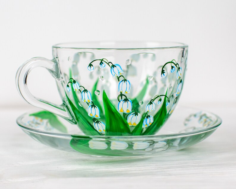 May include: A clear glass teacup and saucer set. The cup and saucer are decorated with hand-painted lily of the valley flowers in white, blue, and green. The cup has a large handle and sits on a matching saucer.