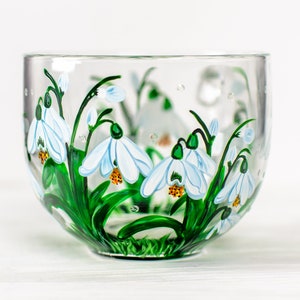 May include: A clear glass mug featuring a hand-painted design of white snowdrop flowers with green stems and leaves. The flowers have yellow centers and are depicted in a cluster around the exterior of the glass, creating a botanical pattern.