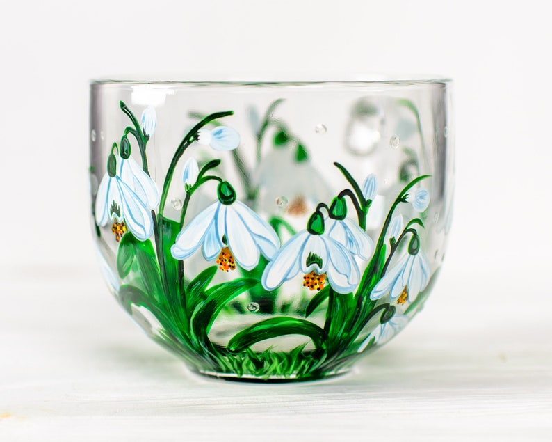 May include: A clear glass mug featuring a hand-painted design of white snowdrop flowers with green stems and leaves. The flowers have yellow centers and are depicted in a cluster around the exterior of the glass, creating a botanical pattern.