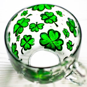 May include: A clear glass mug with a handle, decorated with hand-painted green shamrocks. The shamrocks are of varying sizes and are scattered across the surface of the mug. The mug is sitting on a white surface.