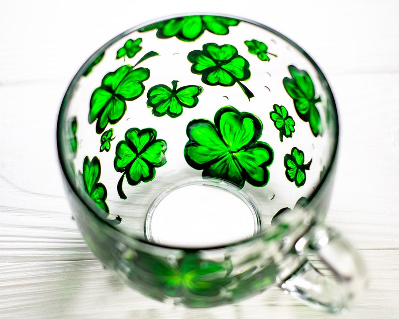 May include: A clear glass mug with a handle, decorated with hand-painted green shamrocks. The shamrocks are of varying sizes and are scattered across the surface of the mug. The mug is sitting on a white surface.