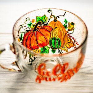 May include: A clear glass mug featuring a hand-painted design of vibrant orange pumpkins, a green-striped gourd, and green leaves. The artwork is rendered in bold, colorful strokes with black outlines, and the mug includes a handle on the left side.