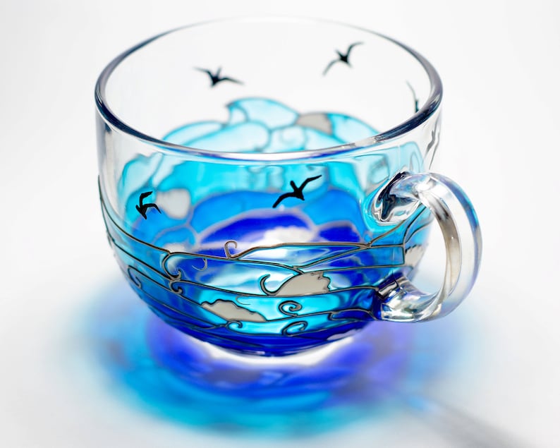 Puede incluir: Una taza de t&eacute; de vidrio transparente con un dise&ntilde;o de oc&eacute;ano pintado. La taza presenta tonos de azul y blanco, que representan olas y gaviotas. El asa es transparente.