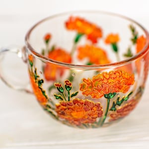 May include: A clear glass teacup with a handle, adorned with hand-painted orange marigold flowers and green stems. The floral design is accented with gold details, creating a decorative and artistic look. The cup is ideal for serving tea or other beverages.
