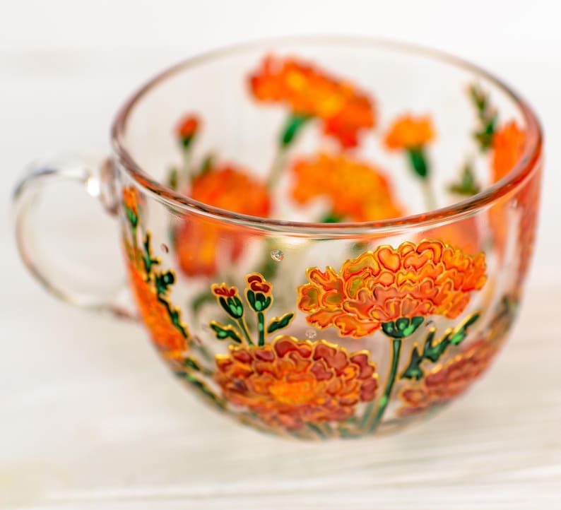May include: A clear glass teacup with a handle, adorned with hand-painted orange marigold flowers and green stems. The floral design is accented with gold details, creating a decorative and artistic look. The cup is ideal for serving tea or other beverages.