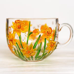 May include: A clear glass teacup with a handle, adorned with hand-painted yellow daffodils and green stems. The floral design includes white accents and is set against a white background. The cup is ideal for tea or coffee.